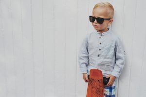 Serious trendy little boy posing with his skateboard wearing a pair of over sized stylish sunglasses borrowed from his father looking towards blank copyspace on a white wall